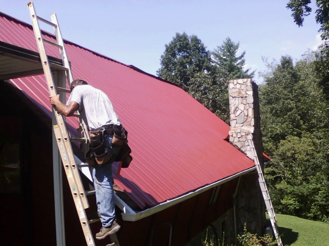 Roofer on a ladder beside a red metal roof and stone chimney — Soffit & Fascia Repair by Roofing Brothers in Westfield Center